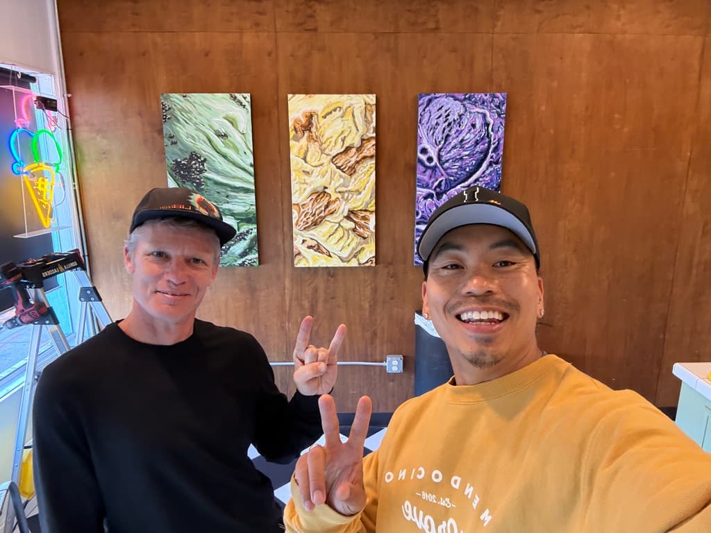 Christopher Bolwyn and a friend smiling in front of the three Ice Cream Trio paintings on The Cookie Bar's wood-paneled wall, with a neon ice-cream-cone sign glowing at left.