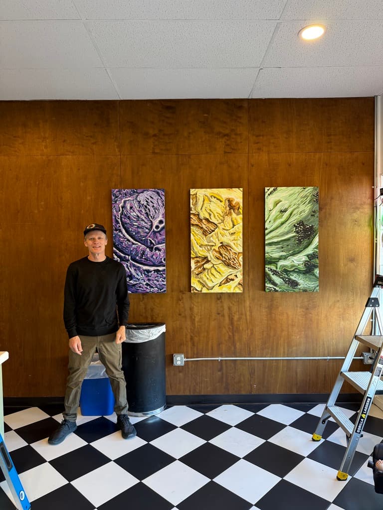 Christopher Bolwyn standing beside the three vertical paintings — Ube, Egg Tart, and Matcha Cookie — on The Cookie Bar's wood-paneled wall, above the black-and-white checkerboard floor, with the install ladder still out.